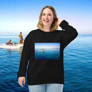 Woman smiling in front of a calm ocean scene with a boat and people, wearing a sweatshirt featuring a photo print of a person on a small boat at sea.