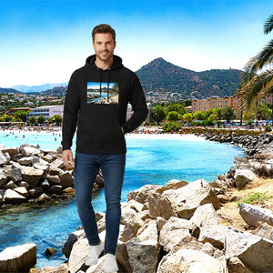 Man in black artistic photo print hoodie standing on rocky shore by a clear blue bay, with mountains and palm trees in the background.