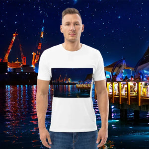 Man wearing white t-shirt with night cityscape art print, standing by illuminated waterfront with cranes and starry sky.